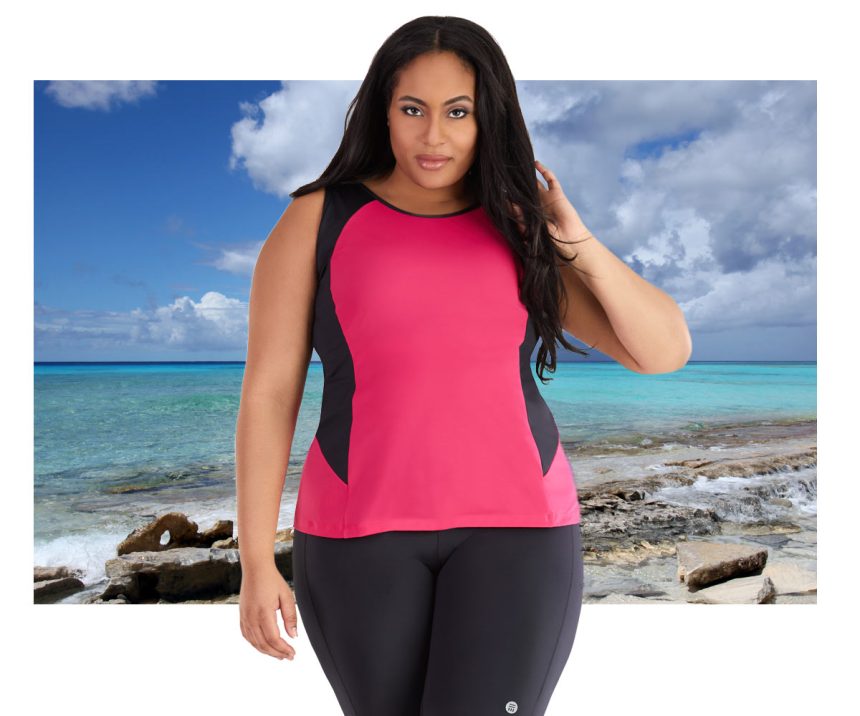 Plus size woman wearing pink and black JunoActive AquaCurveTankini Top and black JunoActive swim shorts. The background is the beach at the ocean.